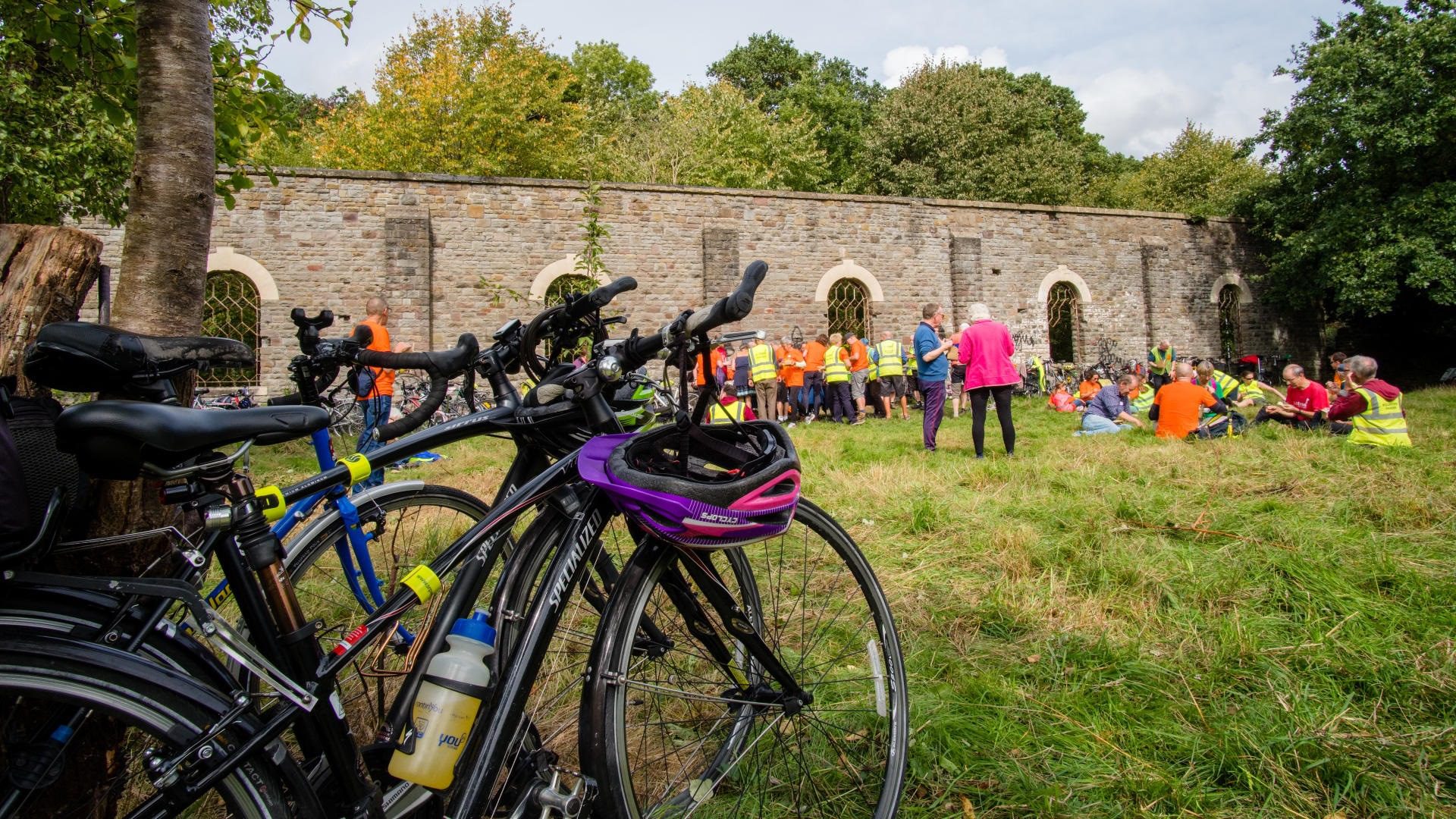 People in high vis jackets sitting in a grassy field in front of an old railway wall with bikes leaning against trees.