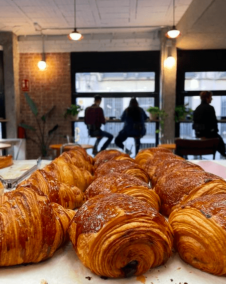A surface filled with croissants and pan au chocolat in front of a window where people sit on high stools.