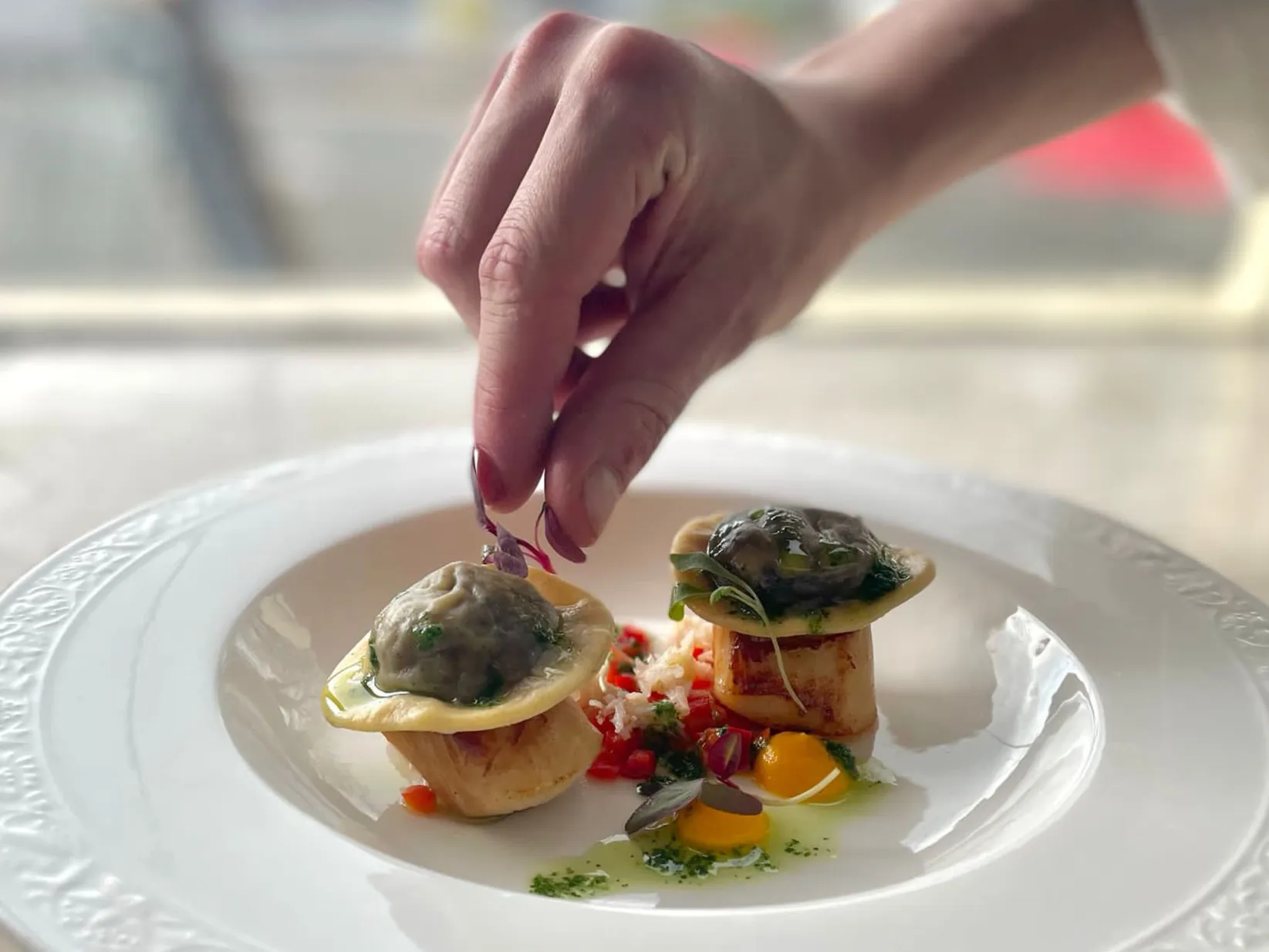 A hand putting micro herbs onto a white plate of food (scallops and tortellini).
