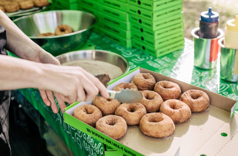 A hand holding tongues hovering over a green box of sugar dusted donuts.