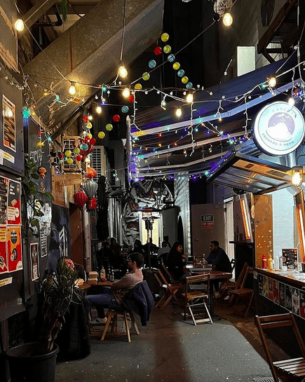 A laneway with people sat on chairs outside and food hatches with fairy lights and lanterns overhead.