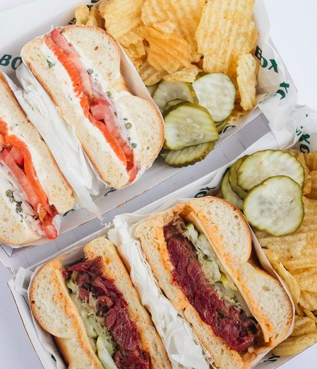 An overhead shot of two boxes of bagels cut open and filled with salmon/ pastrami, with crinkle chips and pickles on the side.