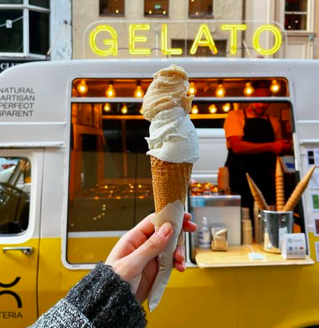 A hand holding up a two scoop cone of gelato in front of a yellow bus which has a neon yellow sign saying 'gelato'.