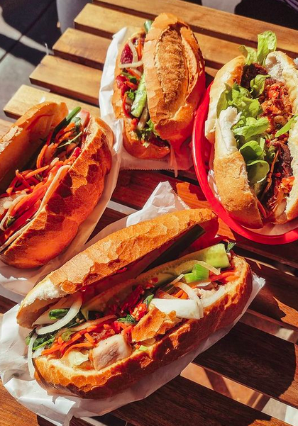 A wooden slatted table with four vietnamese baguettes on top filled with proteins, salad and sauce.