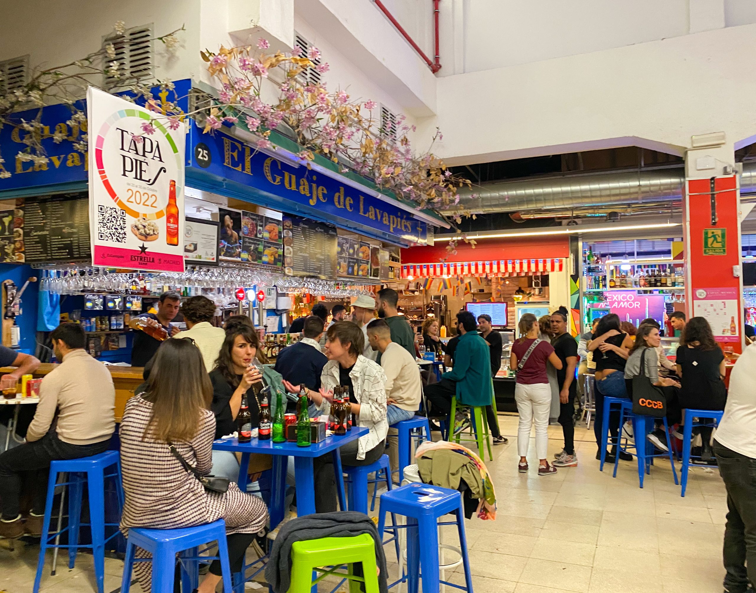 A busy food hall with people queuing at a bar for drinks and other people chatting on high stools clustered around tables.