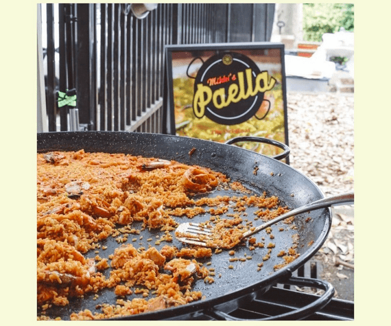 A large paella dish half full of seafood paella with a spatula resting on the pan and a sign saying 'Mikki's paella' in the background.