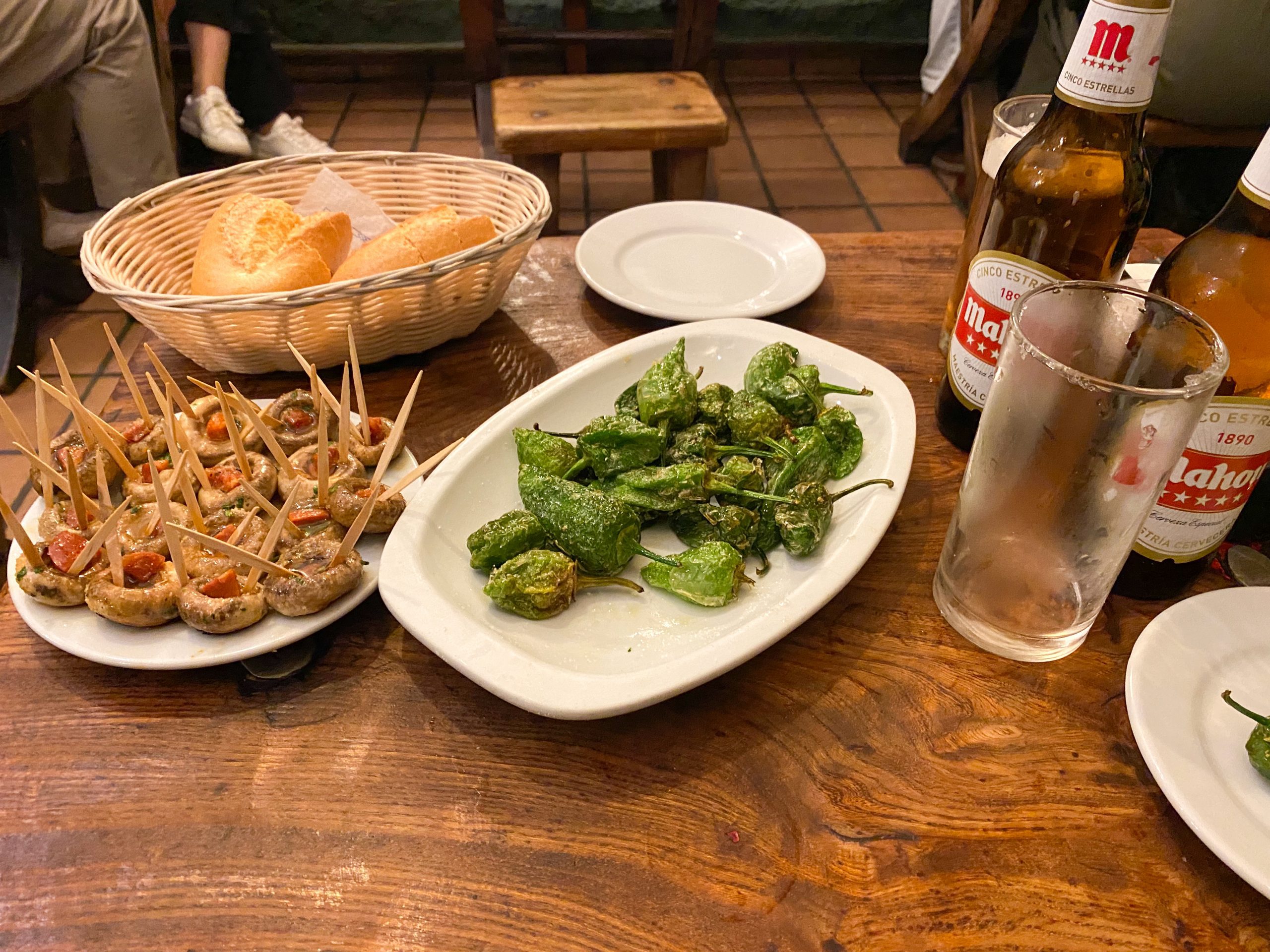 A plate of stuffed button mushrooms and a plate of green salted peppers on a wooden table with some bottles of beer and a bread basket. 