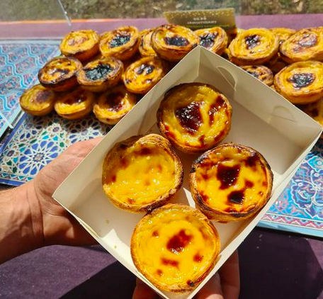 A hand holding a white paper box filled with four portuguese tarts, with a further tray of tarts in the background.