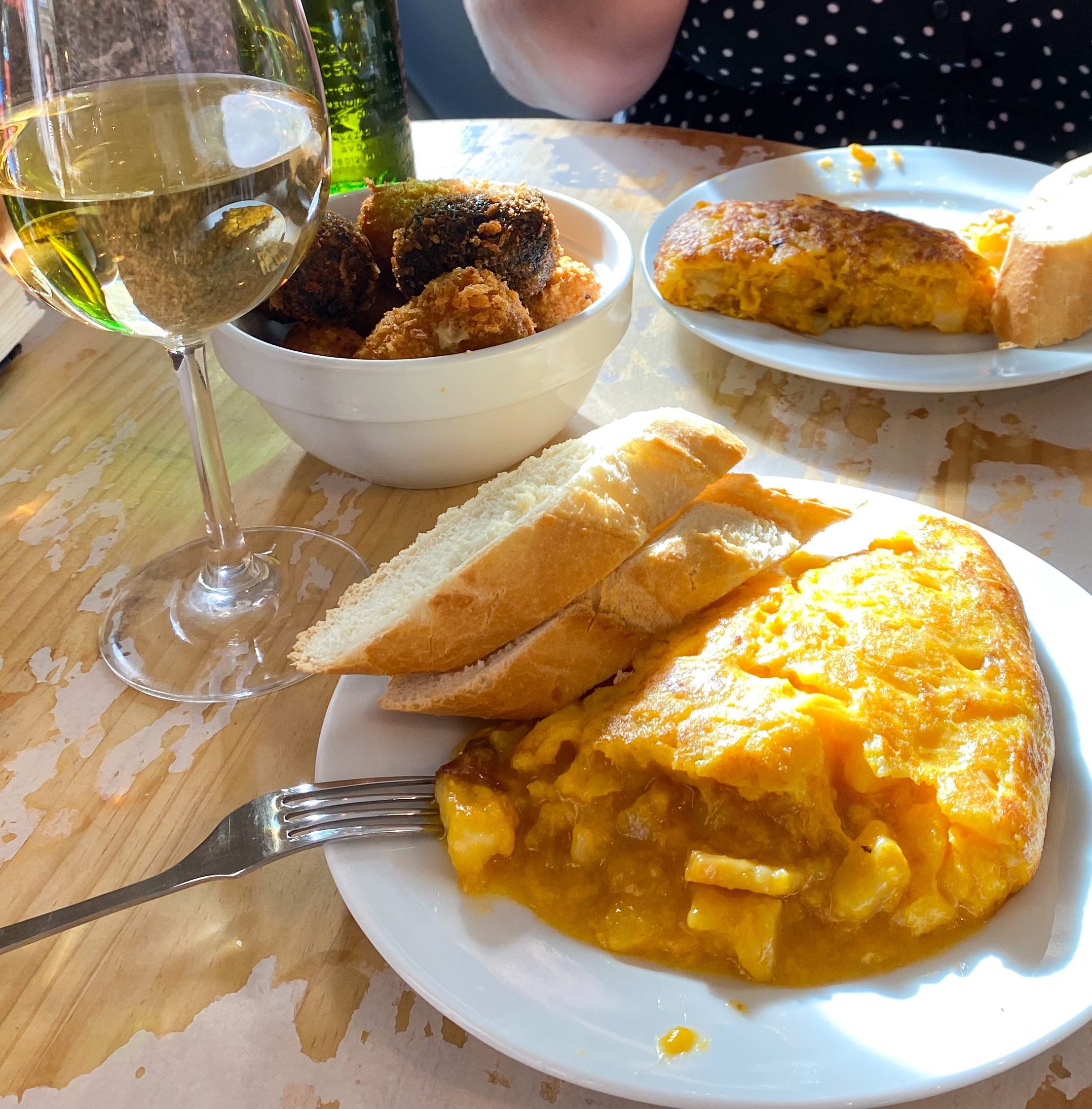 A table with plates of spanish omelette and bread, a bowl of croquettes and some alcoholic drinks.