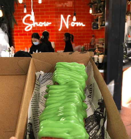A cake covered in green custard in a cardboard takeaway box being held up in front of a neon sign saying 'show nom'.