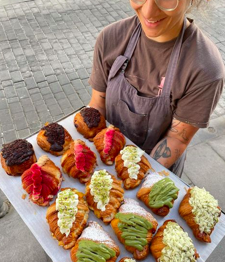 A person in a grey apron holding a tray of stuffed croissants with different coloured cream on top.