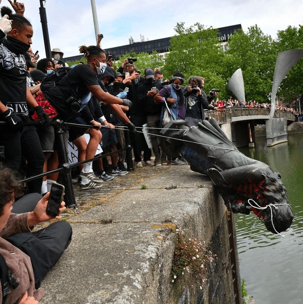 A group of people toppling a black statue into a river using ropes.