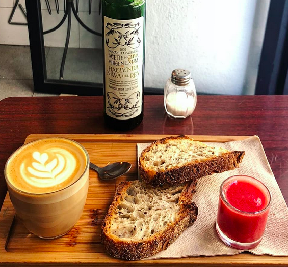 A wooden tray with toasted bread, a small glass of pureed tomato and a coffee with milk, in front of a bottle of olive oil and a salt dispenser.