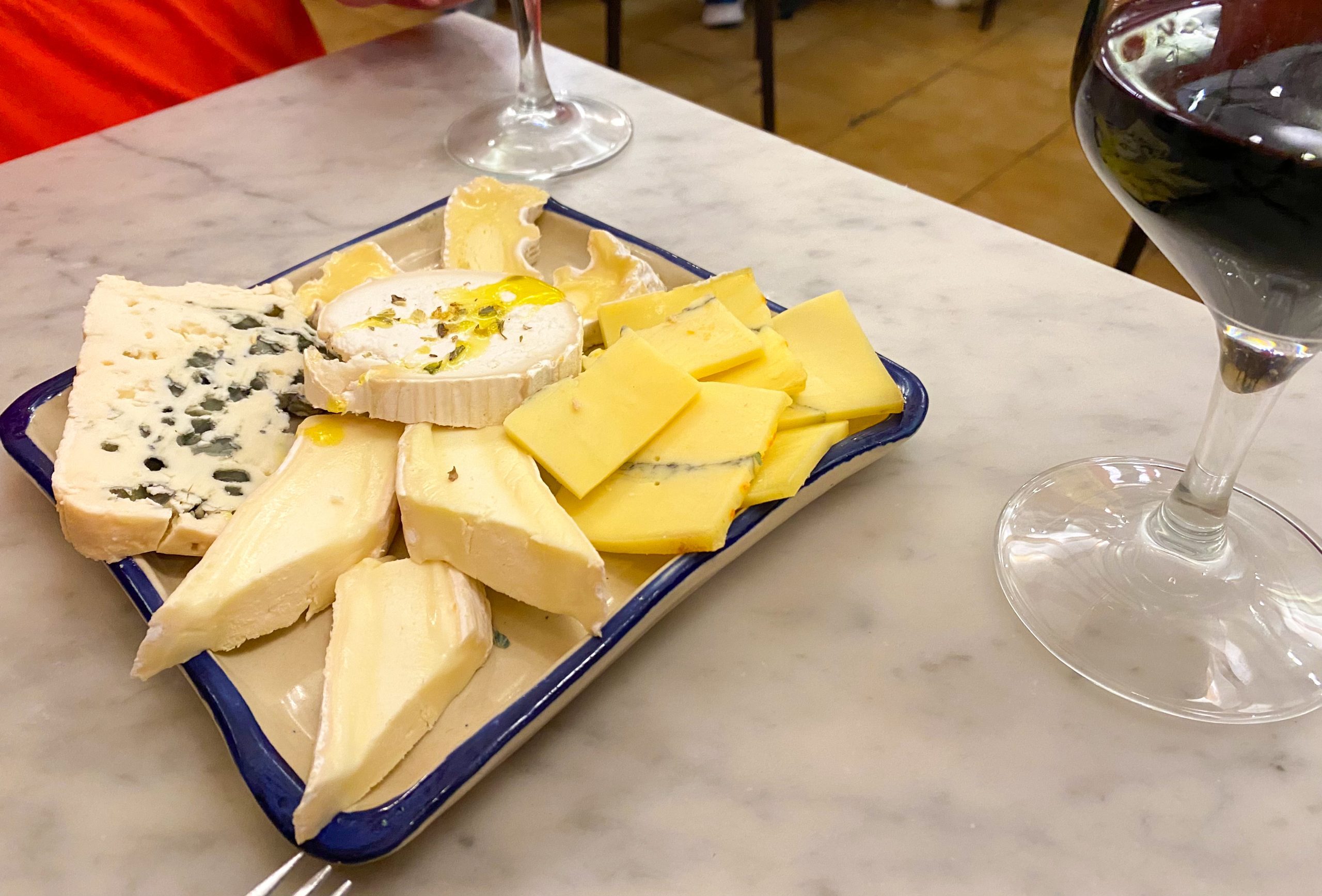 A plate of assorted slices of cheese on a marble table with two glasses of red wine.