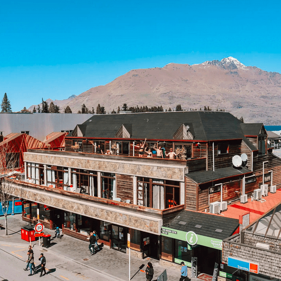 An alpine style building on a roadside with people waving from the upstairs terrace and a mountain in the distance.