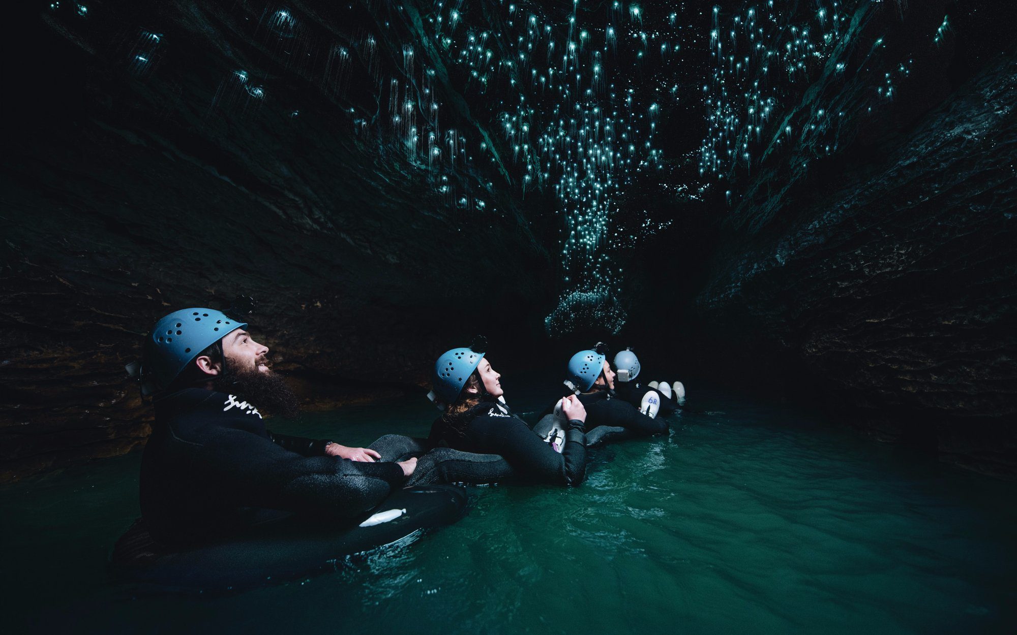 Four people floating in a cave of water in black rubber rings with black wetsuits and blue helmets on looking up at glow worms on the roof.