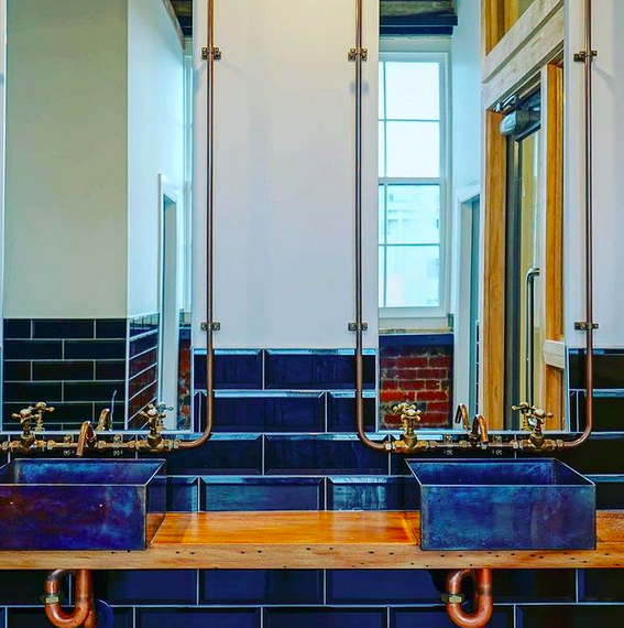 2 bathroom sinks with industrial copper piping and black bowls on top of a wooden shelf.