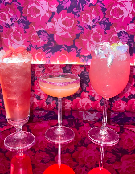 Three pink cocktails on a mirrored table in front of a wall covered in pink floral wallpaper.