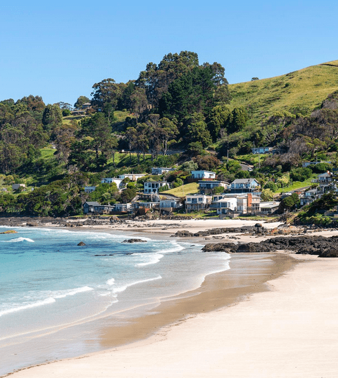 A curved beach with clear blue water and gentle waves with houses and trees on a hill in the backdrop.