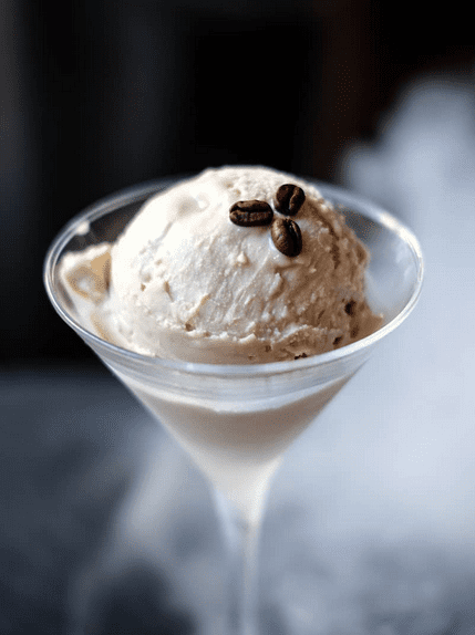 A martini glass filled with cream coloured alcoholic icecream with three coffee beans on top.
