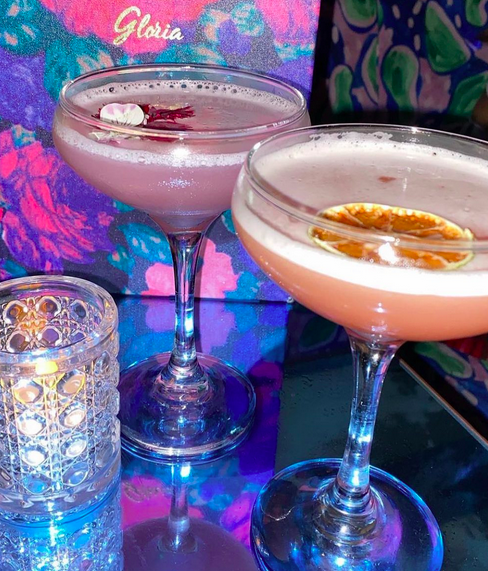 Two martinis on a reflective table next to a candle in a jar and a neon floral menu behind it.