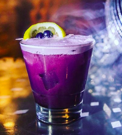 A purple cocktail with foaml a lemon slice and blueberries on top