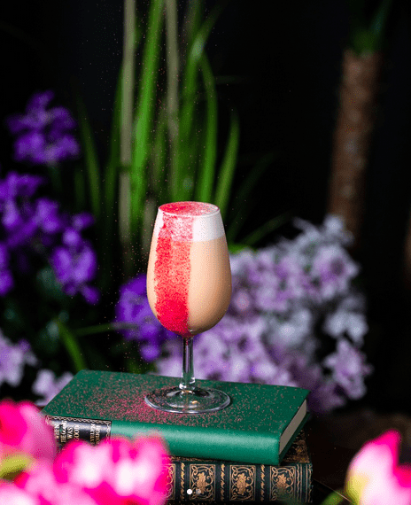 A pink cocktail with foam and pink powder on top of two old bound books with purple flowers in the background.