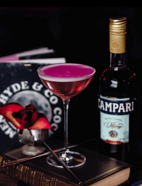 A pink tall cocktail sat on top of an old book with a bottle of campari and a cocktail menu in the background.