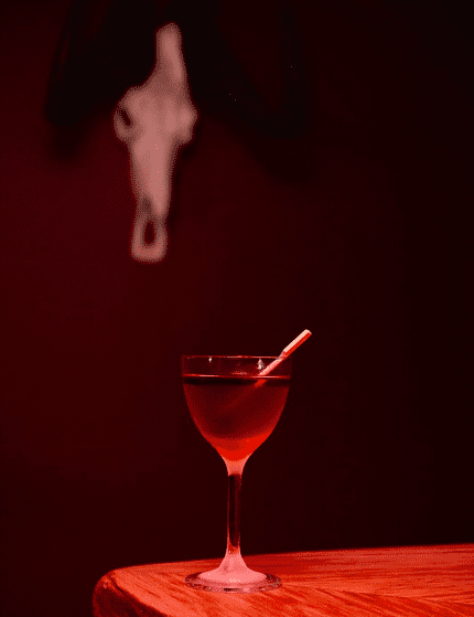 A small martini glass on a wooden surface filled with a red liquid with a wooden stirrer in it.