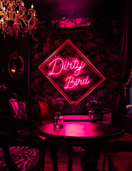 A cocktail bar with a neon sign saying 'Dirty Bird' and tables with vases of flowers on.
