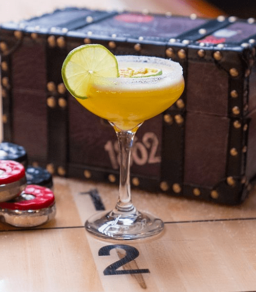 A margarita cocktail garnished with a slice of lime in front of a brown leather chest with shuffleboard pucks next to it.