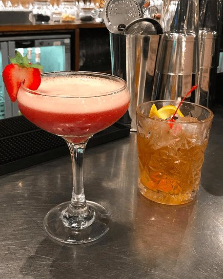 A strawberry cocktail in a martini glass and a brown cocktail in a short glass on a metal bar with a cocktail shaker and a fridge in the background.