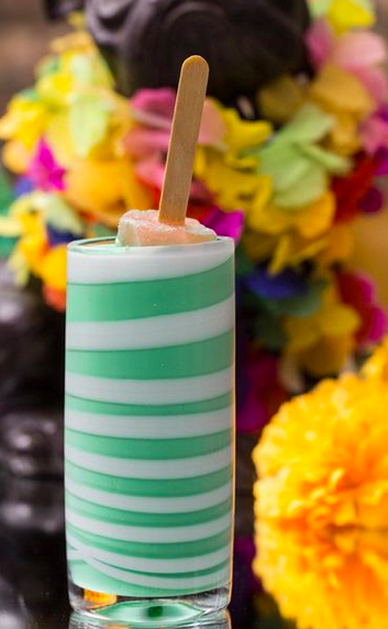 A green and white stripey cocktail with a small ice lolly stuck in the top on a surface with flowers in the background.