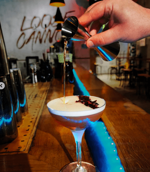 A hand pouring liqueur into a martini glass with foam and edible petals on top.