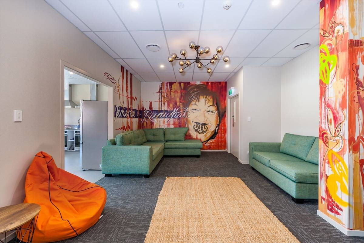 The communal area of a hostel with orange graffiti paintings on the walls, green sofas, an orange bean bag and a beige rug.