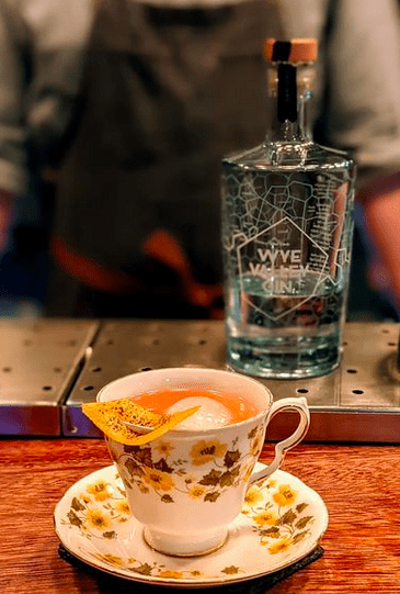 A teacup on a saucer filled with a gin cocktail in front of a bottle of clear gin.