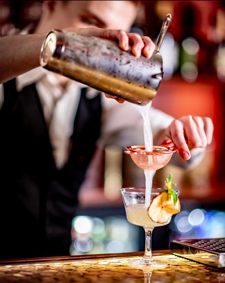A waiter at a bar pouring the contents of a cocktail shaker through a strainer into a champagne glass garnished with an apple.