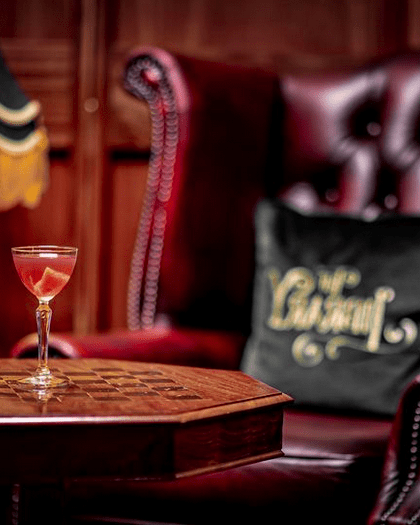 A pink cocktail in a small martini glass on a wooden table in front of a red leather chesterfield chair.