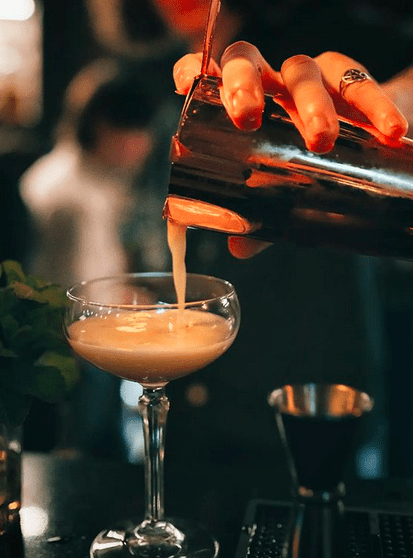 A hand pouring a liquid into a martini glass on a bar surface.