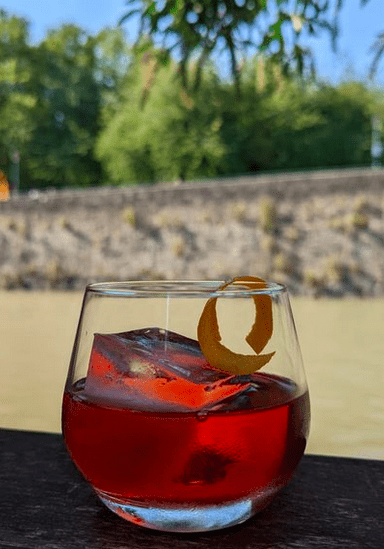 A short glass of negroni on a wooden surface in front of a river with an old stone wall in the background.